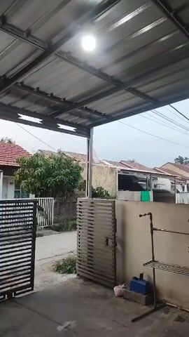 The video shows a covered outdoor area, possibly a carport or patio, with a metal roof and exposed beams. A bright light fixture is attached to the ceiling. In the foreground, there is a grey metal gate on the left and a grey louvered gate on the right, which is slightly ajar. Against the wall on the right, there is a metal drying rack with shelves and some plumbing fixtures. Below the drying rack, there is a blue container and a pink bag. The ground appears to be concrete. Beyond the gates, a street is visible with houses and trees. The sky is overcast, suggesting it may be daytime or dusk. There are no people visible in the video. The overall impression is a residential outdoor space.