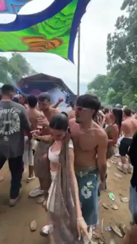 The video shows a group of people dancing at an outdoor festival. A large, colorful canopy hangs overhead. Many of the people are dressed in summer clothing and appear to be having a good time. A DJ booth can be seen in the background, emitting smoke. The ground is covered in dirt and debris. The camera is moving and shaky.
