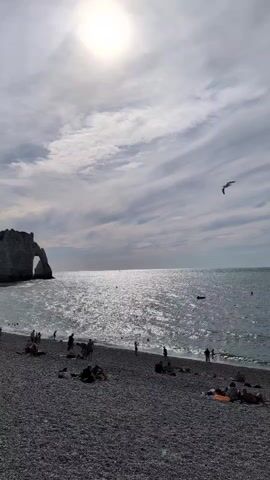 The video shows a wide shot of a pebble beach with people relaxing and walking. In the background, the ocean stretches to the horizon, with the sun reflecting brightly on the water. A natural rock arch formation is visible on the left side of the frame. A small airplane is seen flying in the sky above the water. The sky is partly cloudy, with streaks of clouds. People are scattered across the beach, some sitting, some standing, and some walking towards the water. A few small boats are visible in the distance on the sea. The overall atmosphere appears to be a calm, recreational day at the beach.