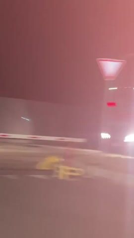 The video captures an outdoor scene at night, likely in Nerubais'ke, Ukraine, given the provided context. The setting appears to be a checkpoint or a toll booth area, with a barrier gate across a paved road. In the foreground, a white barrier with red stripes is visible, and a yellow traffic cone or bollard is on the right.

Several vehicles are present. A red truck with white detailing is seen in the background, and a white passenger van is positioned in front of it. Another white van, similar to the first, is further in the background on the left. A third white van is on the right side of the frame, facing away from the viewer.

A person wearing a blue t-shirt and black shorts with a backpack walks across the paved area in the foreground. The person appears to be moving from left to right. The overall lighting suggests it is nighttime, with artificial lights illuminating the area. There is no visible text in the video. The atmosphere seems neutral, with no discernible strong emotions.