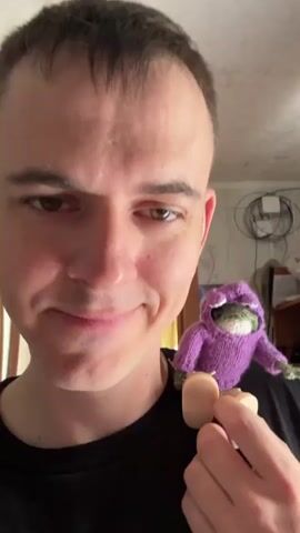 A man with short, dark hair and light skin holds a small, green, knitted frog wearing a purple hoodie in his hand. The frog has light-colored shoes. The man is wearing a black shirt. He smiles slightly and looks at the camera. The background is blurry, but it appears to be a room in a home.