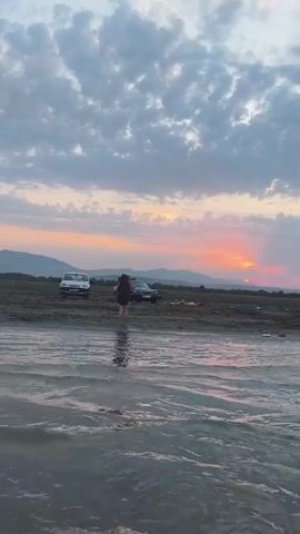 The video shows a wide shot of a body of water with a person standing in it near the shore. The person is wearing dark clothing. There are two cars parked on the dry land behind the person, and a few other objects are scattered on the ground. In the distance, there is a mountain range under a cloudy sky, with the sun setting, casting orange and pink hues across the horizon. The water is calm and reflects the colors of the sky.