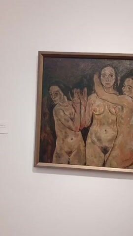 The video shows a framed painting on a white wall in a museum or gallery setting. The painting depicts three nude female figures standing closely together against a dark, indistinct background. The figures are rendered in earthy tones, with visible brushstrokes. The central figure is embraced by the figure on the right. The figure on the left has one hand raised.