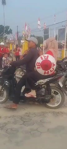 The video captures a street scene with several individuals and vehicles. In the foreground, a person is riding a black motorcycle with a passenger behind them. The passenger is holding a large, circular object with red and white markings, and text that appears to say "TRI" and "CH" in red letters. The rider is wearing a grey cap and a dark shirt, while the passenger is wearing a hijab and a red top. Several other motorcycles are visible, including a white scooter in the middle ground. In the background, a group of people in red shirts and caps are gathered, some standing and some possibly marching or parading. There are also flags visible, including an Indonesian flag. A building with a red tiled roof and yellow walls is present in the background, and some trees and foliage are also visible. The ground appears to be paved with cobblestones or similar material. The overall impression is of a local event or gathering.