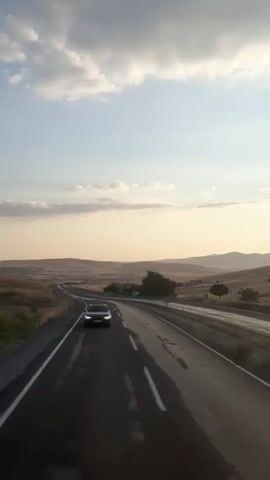 A dark colored car is driving on an asphalt road in a rural landscape during the day. The road is wet and reflects the light. The sky is partly cloudy with a pale blue hue. In the distance, there are rolling hills covered with dry grass and scattered trees. The sun is setting, casting a warm glow on the landscape. The camera is mounted inside a vehicle and is looking forward through the windshield. The car in front is a sedan and its headlights are on.