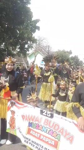 The video shows a street parade with a large group of people in traditional attire. The participants are wearing black tops and skirts made of yellow straw-like material, with headpieces adorned with feathers. Some are carrying staffs. The group appears to be marching down a street lined with buildings and trees. A banner is visible in the foreground with Indonesian text that reads "PAWAI BUDAYA NUSANTARA ADAT PAPUA." There are also spectators on the sides of the street. An Indomaret store sign is visible in the background. The overall atmosphere seems festive.