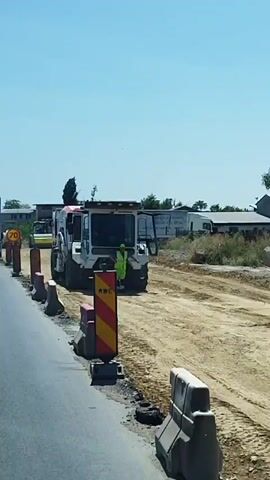 The video shows a construction site on the side of a road. A person in a high-visibility vest is standing near a large piece of construction equipment, possibly a milling machine. There are several trucks and a road roller present at the site. Traffic cones and barriers are set up to direct traffic. The weather appears to be clear and sunny. The video moves from the construction site to a shot of traffic on the road, showing several trucks passing by.