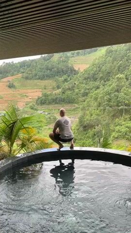 A bald man in a gray shirt and black shorts squats on the edge of a hot tub, looking out at a hilly, green landscape with rice terraces. He points his right index finger upwards.