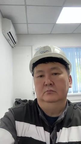 This is a selfie video of a person wearing a white hard hat and a gray and black work uniform. The person is looking directly at the camera with a neutral expression. The background appears to be an office or a work environment, with a window with vertical blinds on the left, a desk with a computer monitor and a printer, and shelves filled with binders and boxes on the right. The ceiling has fluorescent lighting. The video is very short, with no discernible actions or interactions.