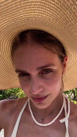 Here's a description of the video:
A young woman is taking a selfie. She is wearing a large, floppy sun hat, a white top, and necklaces. She is standing outside with a beautiful view of the ocean and a tropical coastline in the background. Several boats are visible on the water. She looks at the camera, smiles, and then looks away. A white flower is pinned to her top. The weather appears sunny.