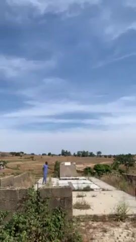 The video shows a man standing on top of a partially destroyed building, gesturing and talking. The sky is blue with wispy clouds. The surrounding landscape appears to be dry, with sparse vegetation and distant buildings or structures. The man is wearing a blue outfit and is the only visible person. The building he stands on seems to be a concrete structure with broken walls and open areas. There are small trees and plants growing around the building.