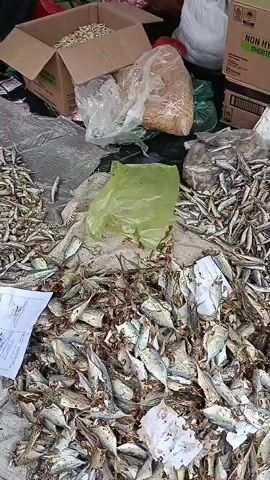 The video shows an outdoor market stall with large piles of dried fish. In the foreground, there is a mound of larger dried fish, some of which are still partially intact. Behind this, there is a pile of smaller dried fish, also spread out on the ground. To the right, several boxes are stacked, one with text that appears to be "NON HYDROGENATED SHORTENING". There is also a green plastic bag and other plastic bags containing what appears to be dried food items like beans or nuts. The lighting suggests it is daytime, and the overall impression is of a busy market selling dried seafood and other goods.