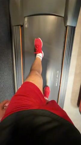A first-person perspective video shows a person running on a treadmill. The person is wearing red shorts, white socks, and red running shoes. The treadmill is gray with orange accents. The background is blurred, suggesting motion. The camera is positioned to capture the runner's legs and the treadmill belt.
