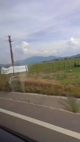A video captured from a vehicle shows a view through a window of a landscape. In the foreground, the side of the road with a white line is visible, along with the edge of the vehicle. Beyond the road, there is a field of grass and some dry vegetation. A tall utility pole with electrical wires stands in the middle of the field. In the background, a mountain range is visible under a cloudy sky. The video is very short, less than five seconds.