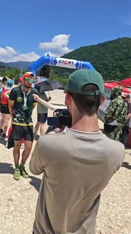 A man wearing a green and yellow shirt, black shorts, green socks, and green trail shoes is being interviewed by a woman wearing a white t-shirt and light brown trousers. The man has a medal around his neck. He is also wearing sunglasses and a cap. The interview is being filmed by a man from behind the camera. The interview takes place in a dirt field under a bright blue sky. In the background, there is a blue inflatable arch with the word "СТАРТ" written on it in white. There are also green mountains in the distance.