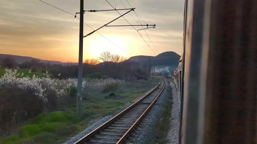 The video shows a view from a train traveling along railroad tracks during a sunset. The sun is low on the horizon, casting a golden glow over the landscape.  The train is moving along a slight curve in the tracks, and the side of the train is visible in the right of the frame. The surrounding area consists of fields, trees, and a small hill in the distance. Overhead power lines are visible. There is some fog or haze in the distance. The scene evokes a sense of travel and peacefulness.