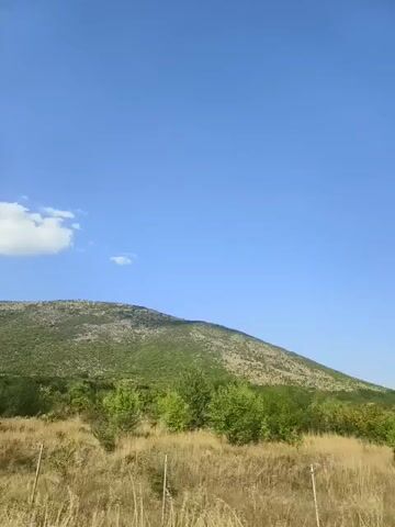 The video shows a landscape with a large hill covered in vegetation under a clear blue sky with a few scattered clouds. In the foreground, there is dry grass and some sparse bushes. A low, weathered concrete wall runs horizontally across the middle of the frame, with some metal posts or fences visible in front of it. The camera appears to be moving, possibly from a vehicle, as the perspective of the foreground shifts slightly. The overall scene suggests a rural or natural environment on a sunny day.