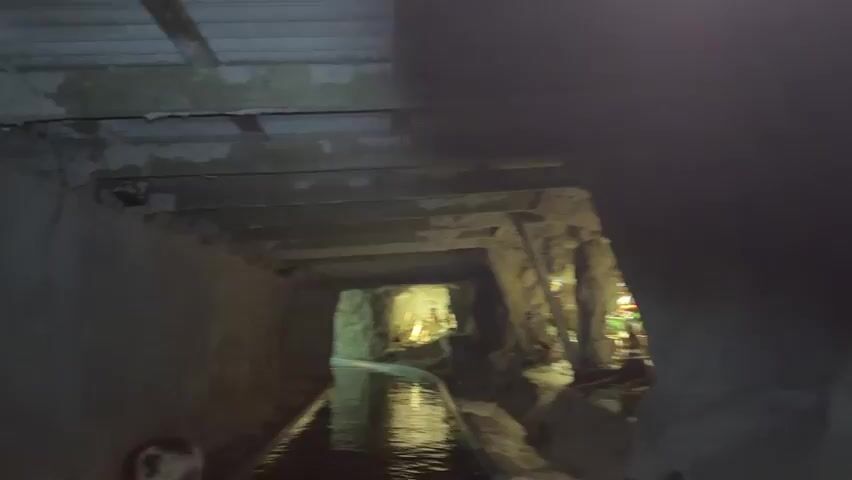 This video shows a first-person view from a boat traveling through a dark tunnel. The tunnel walls appear to be made of rough stone and are supported by wooden beams overhead. The water in the tunnel is dark and reflective, with ripples indicating movement. On the right side of the tunnel, there are several small dioramas with figurines. One diorama features several figures that appear to be miners, dressed in traditional mining attire and engaged in various activities. Another diorama further down the tunnel also contains figurines. The lighting in the tunnel is dim, with spotlights illuminating the dioramas and casting shadows. There are also glimpses of other people in the boat in the foreground.