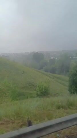 This video shows a view from a vehicle traveling on a road. The weather appears to be overcast or foggy. The landscape consists of rolling green hills, trees, and some vegetation. In the distance, there are some buildings, possibly a village. There are a few dark shapes that could be animals, possibly cows, on the hillside. The road itself has some gravel and dirt. The perspective suggests the camera is positioned to capture the passing scenery through a window.