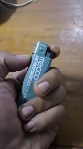 A close-up shot focuses on a hand holding a gray M2000 lighter with a flame igniting. The lighter has hashtags and text printed on its side. The flame flickers, changing color from yellow to blue, indicating a change in the burning process. The background is out of focus, showing a wooden surface and a white wire.