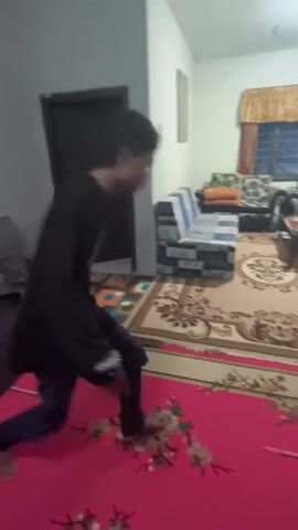 The video shows a person in a black jacket and jeans performing a breakdancing move in a living room. The person starts in a crouched position, then kicks their legs out and spins around on their hands and feet, transitioning to a kneeling position. The living room has a large patterned rug, a coffee table, sofas, and a window with curtains. There is a pink mat on the floor in the foreground. The lighting is provided by an overhead light fixture.