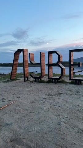 The video shows a set of large wooden letters spelling "UVBEUD" on a sandy beach. Behind the letters, there's a body of water reflecting a sunset. Distant mountains are visible on the horizon. The sky is a mix of blue, orange, and pink hues. There's a stick lying on the sand in the foreground.