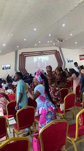 The video depicts an indoor gathering, likely a religious service or event, with rows of red chairs facing a stage area. The ceiling is white with circular light fixtures. In the background, a large screen displays an image of several individuals in academic regalia, possibly graduates. Several people, predominantly women dressed in colorful, traditional African attire, are visible standing and milling around the chairs. One woman in a bright blue and pink dress with a matching headwrap is actively engaged, gesturing with her hands and appearing to speak. A man in a light blue garment stands near her, looking in her direction. Further back, other attendees are seated or standing, some appearing to watch the proceedings on the screen. Another man in a tan tunic is visible on the right side of the frame. The overall atmosphere appears lively and engaged.