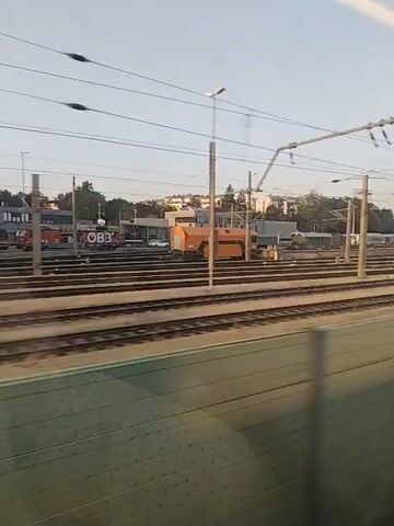 The video captures a view from a moving train, looking out at a train yard under a clear sky. Multiple sets of train tracks run parallel, with overhead electrical lines crisscrossing the scene. In the distance, there are buildings, some appearing to be industrial or maintenance facilities. Several red trains or train cars are visible, as is a white van and a large orange piece of industrial equipment. Fire trucks can also be seen near the buildings. The light suggests it is either early morning or late afternoon. There are no people clearly visible in the frame. The perspective shifts slightly as the train moves, creating a blurred effect on the foreground.
