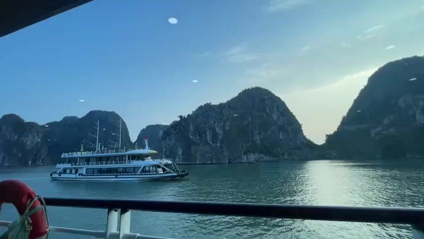 A cruise ship is sailing on a body of water past numerous rocky islands under a clear blue sky. The ship is white with multiple decks and has several windows. The islands are covered in green vegetation and have steep, rocky cliffs. The water is a greenish-blue color. The video appears to be filmed from the deck of another vessel, with a black railing visible in the foreground.