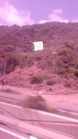 This video shows a view from a vehicle moving along a road. The foreground consists of the road itself, with white lane markings visible. Beyond the road, a low wall with colorful, rectangular segments is present. Further back, a wooded hillside rises, dotted with sparse vegetation and rocks. A large, light-colored rectangular object, possibly a building or structure, is situated on the hillside. A light green, rectangular flag or banner is also visible on the hillside, appearing to be attached to a pole. Power lines run across the scene, supported by utility poles. The sky above is a soft purple hue with scattered clouds, suggesting either sunrise or sunset, or a filter has been applied to the footage. The overall scene is an outdoor, natural environment with some man-made elements. No people are clearly visible, but the context of a moving vehicle suggests the presence of occupants. The atmosphere is calm and the visuals are somewhat hazy, possibly due to the motion or lighting conditions. No text is discernible in the video.