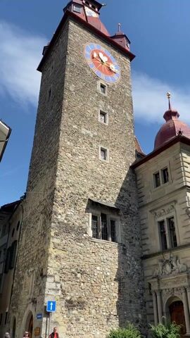 The video pans upwards and across a European cityscape under a bright blue sky with scattered white clouds. The shot begins with a close-up of a tall stone clock tower with a large clock face featuring blue and red details. The tower has a red roof and small windows. To the right of the tower, a building with a red dome-shaped roof is visible. As the camera pans right, the full building is revealed, showing a light-colored stone facade, windows, and a flag with blue and white stripes. The camera continues to pan, revealing other buildings with varied architectural styles, including one with a mural and another with a striped awning in front of it. People are visible in the foreground, including a person wearing a red jacket.