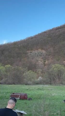 The video shows a rural landscape under a clear blue sky. In the foreground, a man stands reading near a green field with scattered objects like a red barrel and a tire. The view pans to show a small, aged building with a rusty metal door and then moves towards a line of trees against the backdrop of a hillside covered with trees, some still without leaves. The scene suggests early spring.