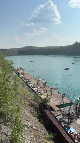 The video shows a turquoise lake surrounded by forested hills. A long wooden pier extends into the water, with many people sunbathing and swimming. Some individuals are seen on paddleboards and small boats. There are also structures on the pier, including a swing set that appears to be used for jumping into the water. The weather is sunny with scattered clouds.