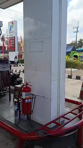 The video pans across a gas station. In the foreground, a white pillar stands prominently, with a red railing in front of it. To the left of the pillar, a fire extinguisher is mounted in a black stand, with a bottle of water nearby. A motorcycle is parked to the left of the pillar. As the camera moves, it shows people and motorcycles at the gas station. One person is seated on a blue and white scooter, facing the camera. Another person, wearing a black shirt and blue shorts, is sitting on a black motorcycle, also facing the camera. A third person is standing next to the black motorcycle, wearing a gray shirt and light-colored pants, with a blue helmet. In the background, there is a Pertamina gas station sign, palm trees, other vehicles, and buildings. The overall scene depicts activity at a busy outdoor gas station.