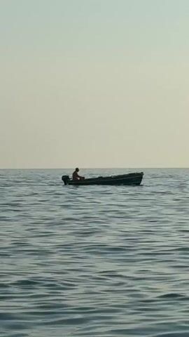A small, dark-colored motorboat with a single person in it is positioned in the center of the frame, on calm, rippling water. The person is seated and appears to be wearing a reddish-orange shirt. The sky is a pale, hazy color, suggesting it is either early morning or late afternoon. To the right of the boat, two buoys or markers are visible, floating on the surface of the water. The boat is moving slowly from left to right.