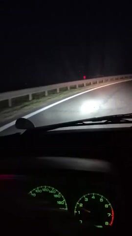 The video shows a first-person perspective from inside a car driving on a road at night. The headlights illuminate the road ahead, which appears to be curved. A guardrail runs along the right side of the road. The dashboard gauges are visible, showing a speedometer and a tachometer, both lit with green light. The speedometer needle is around 120, and the tachometer needle is around 2. The odometer displays "932494." There are red lights visible in the distance, possibly tail lights or marker lights. The overall environment is dark and rural.