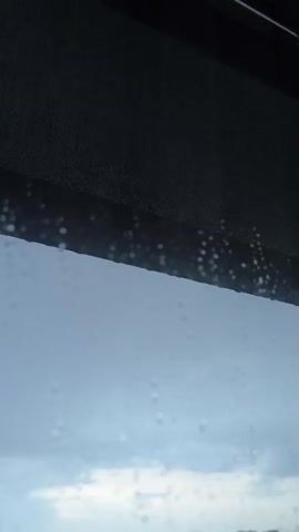 The video shows a close-up view of raindrops on a window. The raindrops are small and scattered across the glass. The background is a blurry landscape of buildings and lights under a cloudy sky. The top of the frame is occupied by a dark, textured object, possibly a window shade or blind. The overall mood is somber and melancholic.