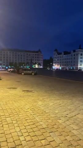 The video opens at ground level, looking out over a cobblestone street or plaza. The sky is a deep twilight blue, indicating either early morning or evening. Several multi-story buildings line the far side of the open space. These buildings appear to be a mix of architectural styles, some with ornate facades and others more modern. Lights are visible in the windows and along the lower levels of the buildings, suggesting that some of them are businesses or residences. Several cars are parked or driving in the distance, and a single red car is visible driving on the right side of the frame. The overall atmosphere is quiet and still, with a sense of urban tranquility.