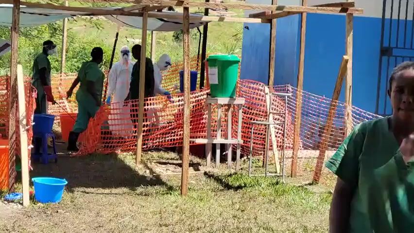 The image captures an outdoor, makeshift health screening or decontamination station in Tchere, Democratic Republic of the Congo, under bright, sunny conditions. The ground is a mix of grass and dirt, with strong shadows indicating midday.

The primary scene features a temporary structure constructed from wooden poles, partially covered by white tarpaulin or fabric, providing shade. An orange safety net barrier delineates an enclosed working area within this structure.

Inside the enclosure, several individuals are present. Two people are dressed in full white personal protective equipment (PPE), including hooded coveralls and masks, suggesting involvement in infection control. Two others wear green scrubs or uniforms; one is seen bending down, possibly interacting with a blue bucket on the ground, while another is partially visible on the far right. A fifth person in darker clothing and a face covering is also within the screened area.

Key objects in the scene include multiple blue buckets on the ground, and a prominent green water dispenser or handwashing station mounted on a wooden stand. These items, along with the PPE, suggest hygiene and decontamination protocols are being observed. In the background, a blue building with a metal gate is visible. The overall setup indicates an organized response or checkpoint related to public health. There is no visible text on any objects.