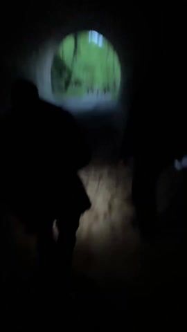 The video shows two people walking from within a dark tunnel toward the exit, where a group of people are visible in the daylight. The tunnel walls appear to be made of stone. The people in the distance are walking away from the tunnel. The surrounding environment outside the tunnel is green with trees.