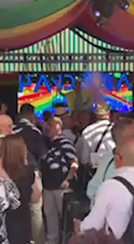 This video depicts a vibrant and festive event taking place in Jerez de la Frontera, likely indoors or under a large awning. The setting is adorned with a colorful, striped ceiling in hues of red, green, yellow, blue, orange, and brown, creating an energetic ambiance reminiscent of a fairground tent or a celebratory venue.

In the background, a prominent digital screen displays animated text that appears to read "PANDORA" or "PANDORA'S," rendered in bright, rainbow colors. This colorful motif, especially the rainbow spectrum, often symbolizes LGBTQ+ pride and suggests an inclusive and celebratory environment.

A diverse crowd of adults is gathered, moving and dancing to upbeat music, indicating an active social event or party. People are dressed casually, with various individuals seen from different angles throughout the short clip. Notable details include a person in a white shirt and dark backpack, and another in a dark top with white stripes.

From the midpoint of the video, the camera pans slightly to the right, revealing more attendees. A woman in a white sleeveless top and a light-colored skirt, featuring a distinctive red accessory in her hair, becomes more visible. Several people are holding small, rectangular objects in blue and pink, possibly balloons, signs, or decorative paddles, some appearing to have glitter, further enhancing the festive mood. The overall activity involves lively social interaction and dancing, with participants appearing to enjoy the event. The lighting is dominated by the bright screen and ambient light, suggesting an evening celebration or a well-lit indoor gathering.