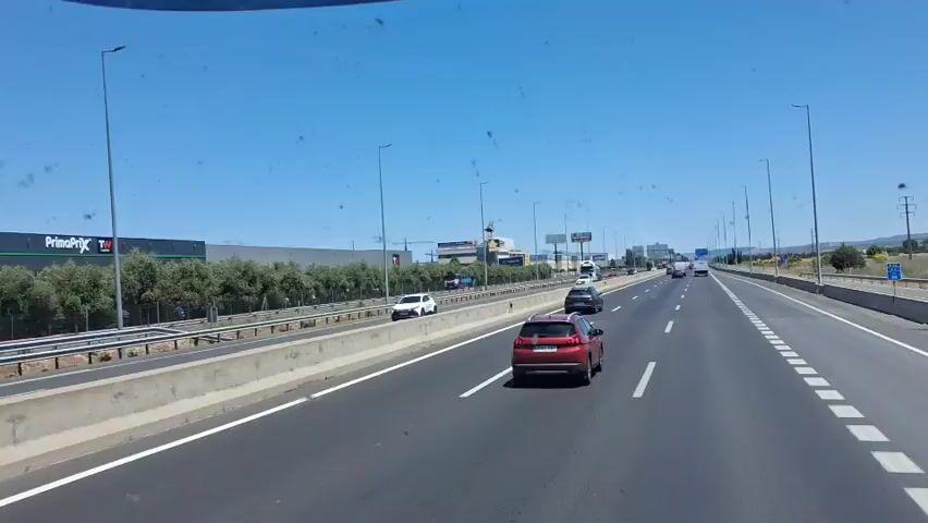 The video shows a car driving on a highway during a sunny day. There are several other cars on the road, and a large building with several signs and a large figure of a cartoon character on its roof can be seen in the distance. The building has signs for "Klaper" and "Logicor". The camera is inside the car, looking out the windshield.