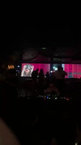 The video shows a dark interior space with multiple people. The focal point of the video is a DJ booth setup, with screens displaying imagery. People appear to be dancing or socializing, and the overall environment suggests a nightclub or party setting. The lighting is dim, but a single spotlight illuminates a section of the crowd at one point. Some individuals appear to be moving around and others are standing still, observing the scene.