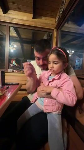 A man sits in a wooden-paneled room, holding a young girl on his lap. The man wears a light-colored t-shirt and has a short, dark beard. He is pointing toward something out of frame. The girl, who is around 4-5 years old, wears a pink denim jacket and a pink headband. She has a serious expression. A table with a cell phone and other items sits to the left of the man. There are windows behind them showing an outdoor scene and a fireplace. Music is playing in the background.