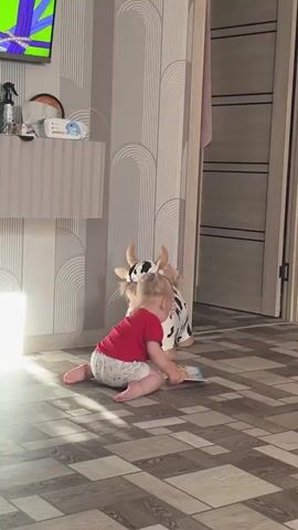 A young child with blonde hair in pigtails, wearing a red t-shirt and patterned shorts, is kneeling on a wooden floor. The child is facing away from the camera, towards a wall with striped wallpaper featuring a repeating arch pattern. Sunlight streams in from the left, illuminating the child and creating a bright spot on the floor. To the child's right is a stuffed toy cow with black and white spots. In the background, a television screen shows a colorful image, and a modern shelf unit holds various items including plants and spray bottles. A doorway is visible on the right side of the frame. The child appears to be interacting with an object near the wall, possibly a light switch or outlet. The scene suggests a domestic indoor setting, likely a living room, during daylight hours given the strong sunlight. The child's posture and focus indicate engagement with their surroundings, and the overall atmosphere seems calm and ordinary.