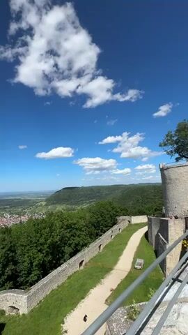 The video shows a panoramic view from a castle wall. The sky is blue with scattered white clouds. Below the castle wall, there is a path with grass on either side. In the distance, there is a town nestled in a valley, surrounded by rolling green hills and trees. The sun is shining, and the overall atmosphere is peaceful and scenic. There are no people visible in the video.