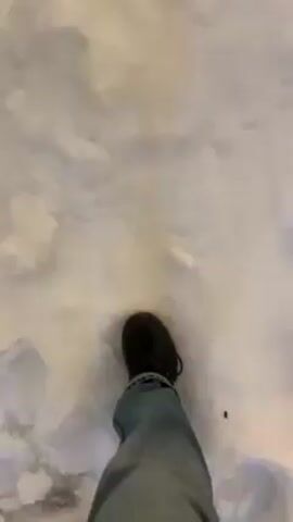 The video shows a first-person perspective of a person walking in snow. The person is wearing blue jeans and black shoes. The snow is mostly white, with some shadows and variations in texture. The person takes several steps, leaving footprints in the snow.