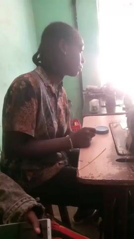 The video shows two people in a room with green walls. One person is seated at a wooden table with a sewing machine. They are wearing a floral patterned shirt. The second person is sitting next to them, wearing a denim jacket over a brightly colored top, and they are holding a smartphone. There is a window with bright light shining through. Music is playing in the background.