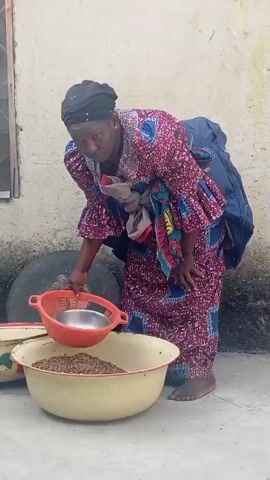 A woman in a patterned dress and headscarf, carrying a baby on her back, lifts a large yellow basin filled with grains. She stands outside a building with a window, tire, and other containers. She lifts the basin and carries it towards a doorway covered with a curtain.