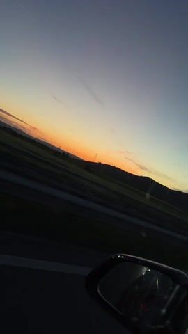 The video shows a view from inside a car at sunset. The sky is a gradient of purple, blue, and orange. In the distance, there are hills and a transmission tower. The side mirror of the car is visible in the foreground, reflecting the interior of the car. The road is visible below the hills, with dark lines indicating lanes of traffic. The overall mood is peaceful and serene.
